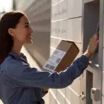A woman holding a small mail parcel using a sunlight readable monitor at a self-service mail kiosk.