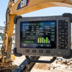 Close-up of a digital monitoring screen mounted on a Caterpillar excavator at a construction site, displaying engine and hydraulic system data while workers operate in the background.
